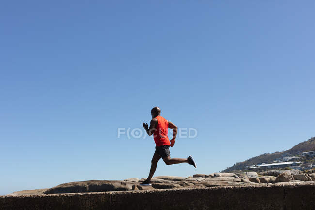 Fit senior african american man exercising running on rocky coastal path. healthy retirement outdoor fitness lifestyle. — Stock Photo