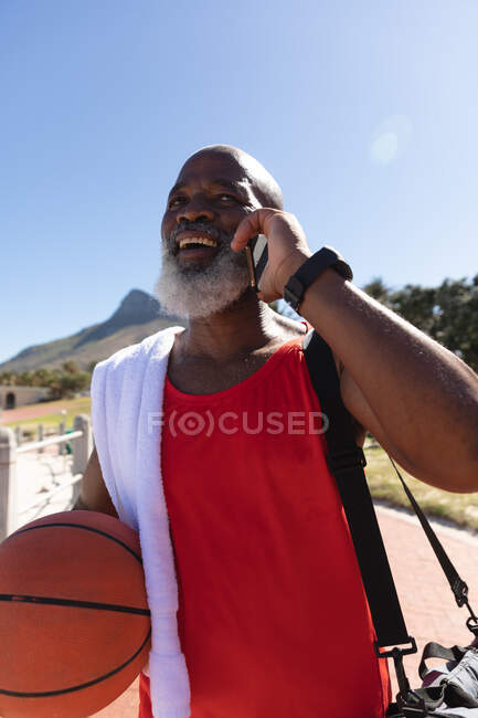 Fit senior homme afro-américain tenant basket parler sur smartphone souriant au soleil. saine retraite technologie communication extérieur forme physique mode de vie. — Photo de stock