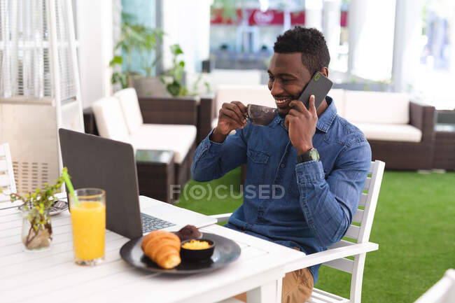 Hombre afroamericano sentado en un café hablando con un smartphone y tomando café. hombre de negocios en la salida en la ciudad. - foto de stock