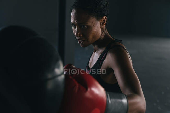 African american woman wearing boxing gloves punching boxing bag in an empty urban building. urban fitness healthy lifestyle. — Stock Photo