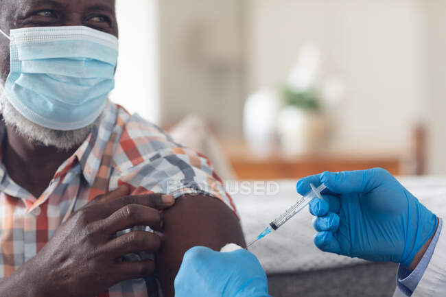 Senior caucasian male doctor vaccinating male patient both wearing face mask at home. healthcare hygiene protection during coronavirus covid 19 pandemic. — Stock Photo