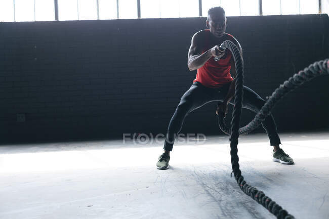 Homem afro-americano vestindo roupas esportivas lutando contra cordas em um prédio urbano vazio. fitness urbano estilo de vida saudável. — Fotografia de Stock