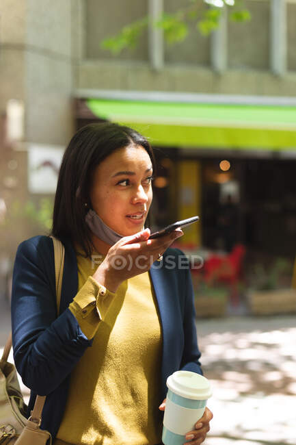 Mujer afroamericana con mascarilla bajada hablando en smartphone en la calle. estilo de vida durante la pandemia de coronavirus covid 19. - foto de stock