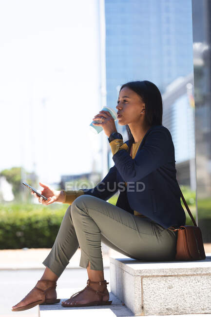 Africano mulher americana com smartphone beber café enquanto sentado no parque corporativo. estilo de vida que vive durante o coronavírus covid 19 pandemia. — Fotografia de Stock
