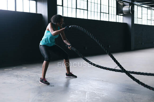 Donna afroamericana che indossa vestiti sportivi che combattono corde in un edificio urbano vuoto. fitness urbano stile di vita sano. — Foto stock