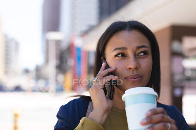 Femme afro-américaine avec tasse de café parlant sur smartphone dans la rue. mode de vie vivant pendant la covie coronavirus 19 pandémie. — Photo de stock