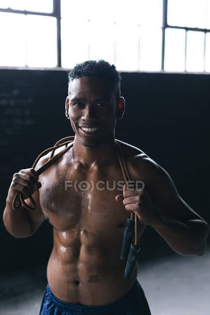 Portrait d'un homme afro-américain ayant une corde à sauter sur ses épaules dans un bâtiment urbain vide. regardant la caméra et souriant. forme physique urbaine mode de vie sain. — Photo de stock