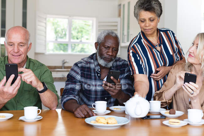 Casais seniores caucasianos e afro-americanos sentados à mesa bebendo chá usando smartphone em casa. sênior aposentadoria estilo de vida amigos socialização. — Fotografia de Stock