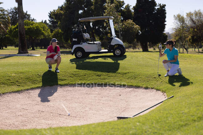 Two caucasian women playing golf squatting down beside bunker talking. sport leisure hobbies golf healthy outdoor lifestyle. — Stock Photo