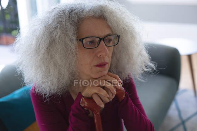 Porträt einer älteren kaukasischen Frau, die im Wohnzimmer sitzt und sich auf einen Gehstock stützt. Während der Quarantäne zu Hause bleiben und sich selbst isolieren. — Stockfoto