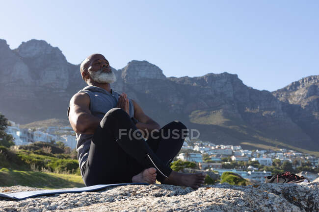 Fit senior african american man sitting on rock by mountain meditating. healthy retirement outdoor fitness lifestyle. — Stock Photo