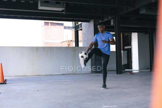Un Afro-Américain faisant des tours avec un ballon de foot dans un immeuble urbain vide. forme physique urbaine mode de vie sain. — Photo de stock