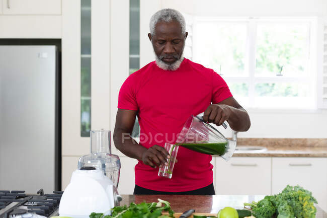 Hombre afroamericano mayor vertiendo fruta y verdura bebida saludable en un vaso. bienestar de la aptitud física en el hogar de ancianos. - foto de stock