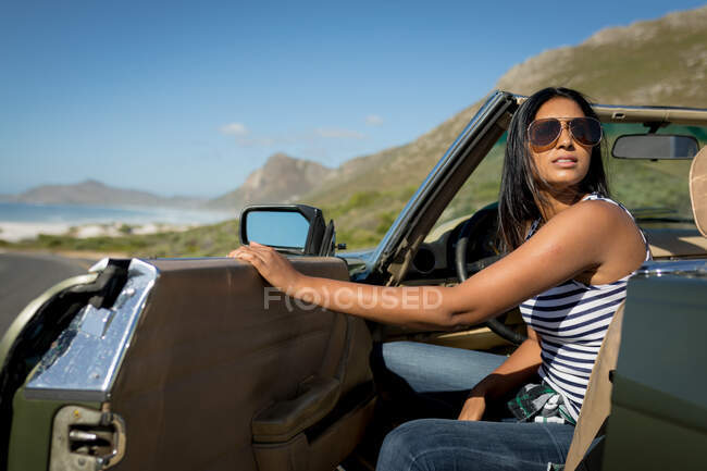 Mujer de raza mixta en día soleado sentado en el coche descapotable. Viaje de verano por carretera en una carretera rural junto a la costa. - foto de stock