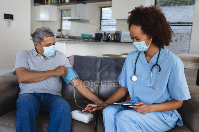 Senior Mixed Race Mann mit Ärztin zu Besuch zu Hause mit Gesichtsmasken Blutdruck messen. Altenpflege zu Hause während Quarantäne-Sperrung. — Stockfoto