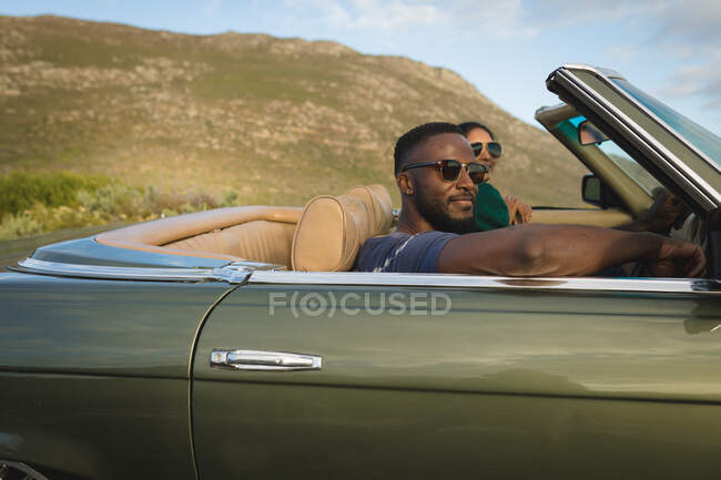 Diverse couple driving on sunny day in convertible car and smiling. Summer road trip on a country highway by the coast. — Stock Photo