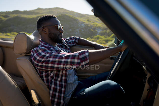 Africano americano dirigindo em dia ensolarado em carro conversível segurando roda motriz. viagem de estrada de verão em uma estrada rural pela costa. — Fotografia de Stock
