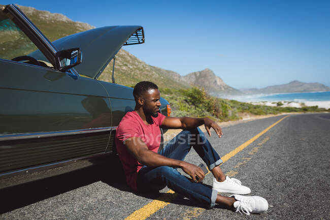 Африканский американец со смартфоном сидит на дороге рядом со сломанной машиной с открытым капотом. летняя поездка по проселочной дороге на побережье. — стоковое фото
