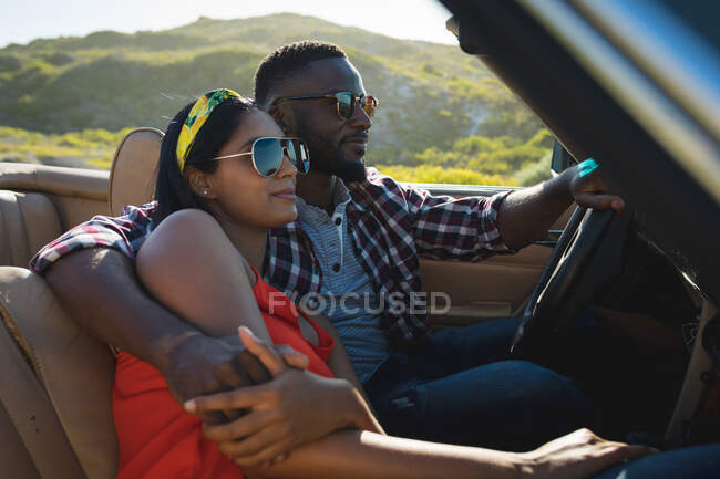 Diverse couple driving on sunny day in convertible car embracing and smiling. Summer road trip on a country highway by the coast. — Stock Photo