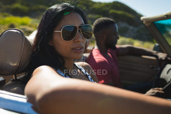 Diverse couple driving on sunny day in convertible car talking and smiling. Summer road trip on a country highway by the coast. — Stock Photo