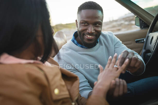 Diverse couple sitting in convertible car man is putting ring on woman's hand. Summer road trip on a country highway by the coast. — Stock Photo