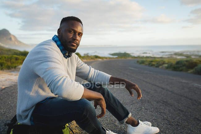 Homem afro-americano sentado ao pé da estrada na mochila. Verão viaja em uma estrada rural pela costa. — Fotografia de Stock