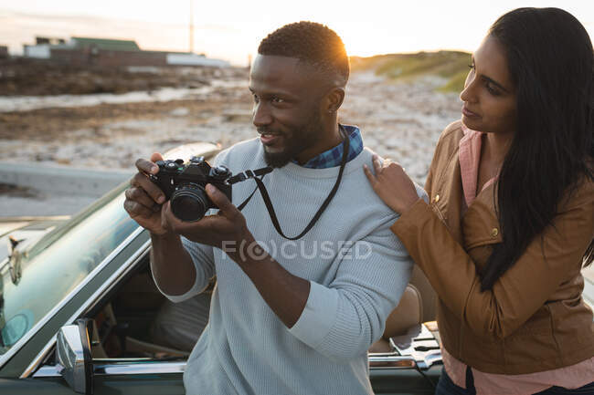 Diverse couple standing by a convertible car and taking photos with camera. summer road trip on a country highway by the coast. — Stock Photo