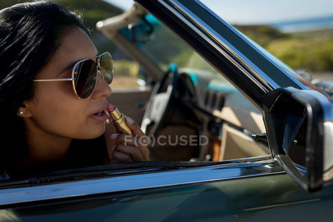 Mixed race woman on sunny day sitting in convertible car putting lipstick on. summer road trip on a country highway by the coast. — Stock Photo