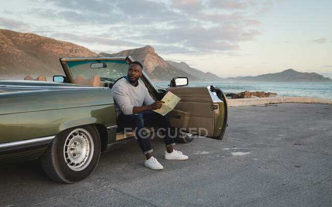 Hombre afroamericano sentado en un coche descapotable y mirando el mapa. Viaje de verano por carretera en una carretera rural junto a la costa. - foto de stock