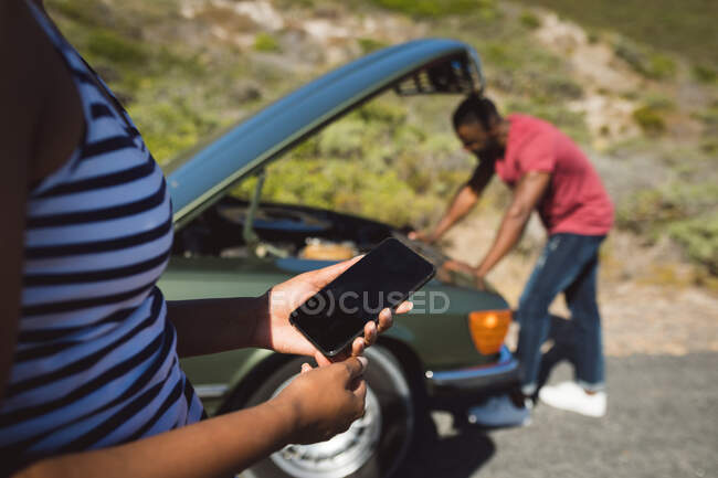 Femme de course mixte utilisant smartphone debout sur la route à côté de la voiture en panne avec capot ouvert. Un Afro-Américain essaie de réparer le moteur.. — Photo de stock