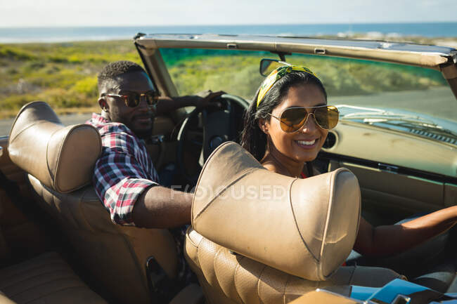 Diverse couple driving on sunny day in convertible car looking at camera and smiling. summer road trip on a country highway by the coast. — Stock Photo