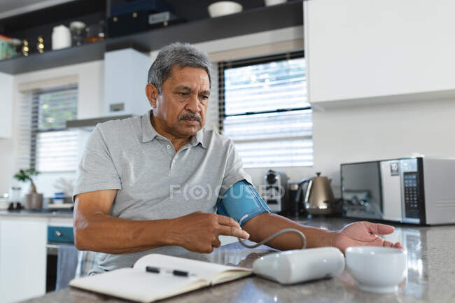 Homme de race mixte senior à la maison prenant sa tension artérielle dans la cuisine. soins de santé primaires à domicile pendant le confinement en quarantaine. — Photo de stock