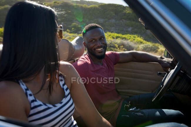 Diverse couple driving on sunny day in convertible car looking at each other and smiling. Summer road trip on a country highway by the coast. — Stock Photo