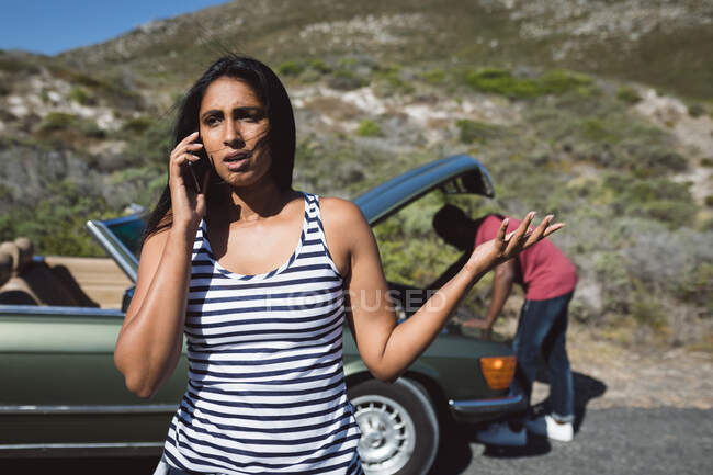 Mujer de raza mixta hablando con un teléfono inteligente de pie en la carretera junto a un coche averiado con capó abierto. hombre afroamericano está tratando de arreglar el motor. - foto de stock