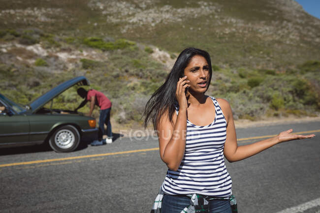 Смешанная расовая женщина разговаривает с помощью смартфона, стоящего на дороге рядом со сломанной машиной с открытым капотом. Африканский американец пытается починить двигатель. — стоковое фото