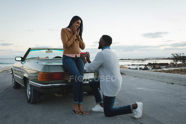 Diverse couple standing by convertible car man is proposing to woman. summer road trip on a country highway by the coast. — Stock Photo