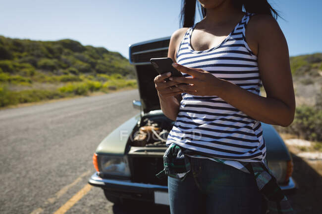 Смешанная гонщица с помощью смартфона стоит на дороге рядом со сломанной машиной с открытым капотом. летняя поездка по проселочной дороге на побережье. — стоковое фото