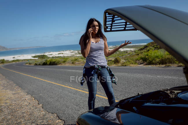 Mulher de raça mista falando usando smartphone em pé na estrada ao lado de carro quebrado com capota aberta. viagem de estrada de verão em uma estrada rural pela costa. — Fotografia de Stock