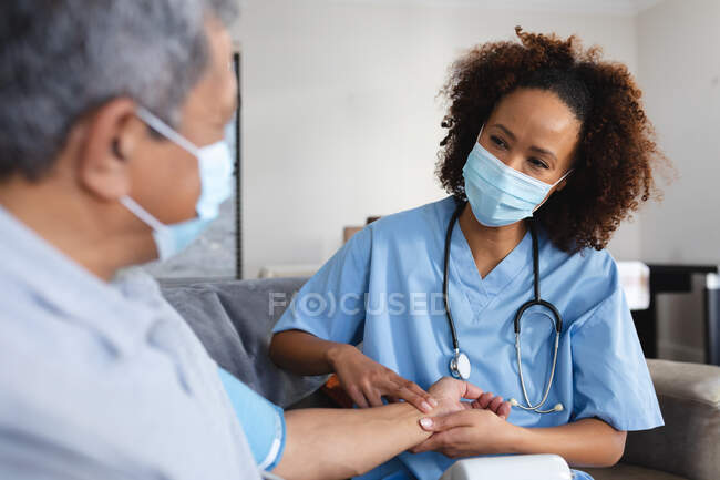 Senior mixed race man with female doctor home visiting wearing face masks taking pulse. senior healthcare at home during quarantine lockdown. — Stock Photo