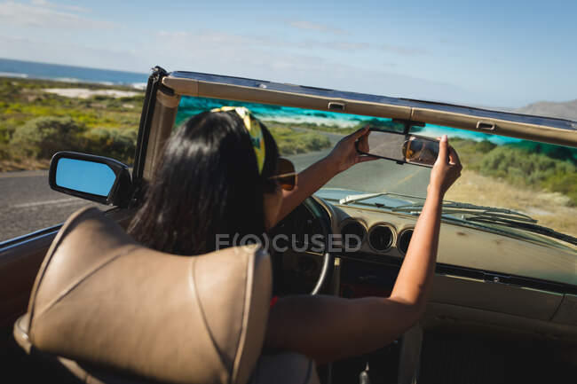 Femme de course mixte conduisant un jour ensoleillé en voiture convertible prenant un selfie. Road trip estival sur une autoroute de campagne au bord de la côte. — Photo de stock