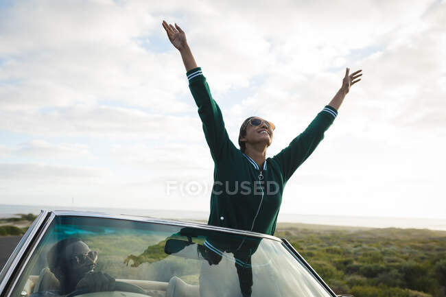 Diversa pareja conduciendo en día soleado en coche convertible mujer está de pie y sosteniendo sus manos hacia arriba. Viaje de verano por carretera en una carretera rural junto a la costa. - foto de stock