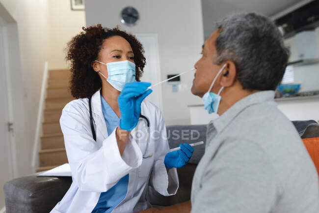 Hombre de raza mixta sénior con médico femenino en casa visitando usando máscaras faciales tomando una prueba de hisopo. salud e higiene durante la pandemia del coronavirus covid 19. - foto de stock