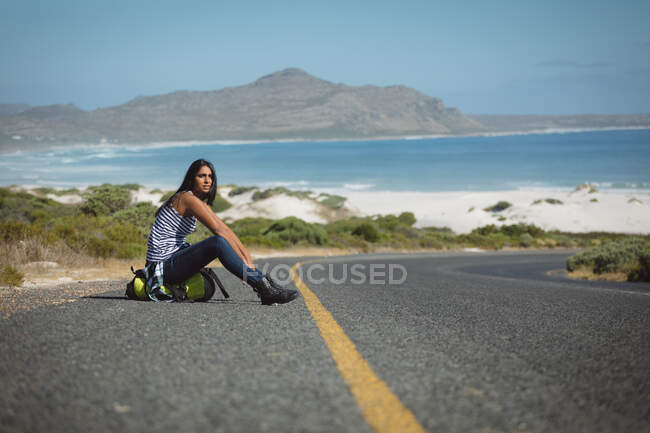 Mujer de raza mixta sentada junto a la carretera y haciendo autostop. verano viaja en una carretera rural por la costa. - foto de stock