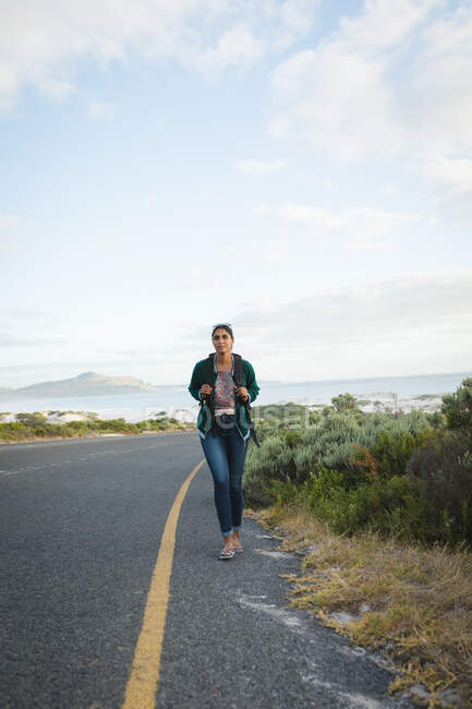 Mujer de raza mixta con una mochila y senderismo en las montañas por la costa. El verano viaja por una carretera rural junto a la costa. - foto de stock