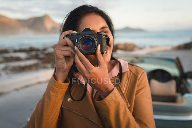 Mixed race woman standing by convertible car and taking photos with camera. Summer road trip on a country highway by the coast. — Stock Photo