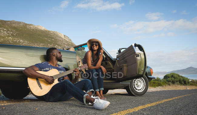 Diversa pareja tomando un descanso en carretera en el día soleado junto a un coche convertible el hombre tocando la guitarra. viaje de verano por carretera en un camino rural junto a la costa. - foto de stock