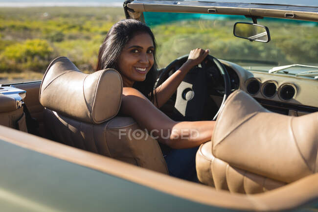 Femme de course mixte conduisant un jour ensoleillé dans une voiture convertible tenant le volant et souriant. Road trip estival sur une autoroute de campagne au bord de la côte. — Photo de stock