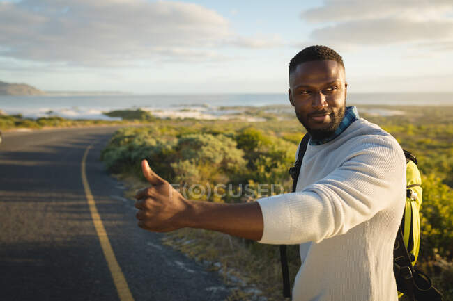 Afroamerikaner steht am Straßenrand und trampt. Sommerreisen auf einer Landstraße an der Küste. — Stockfoto