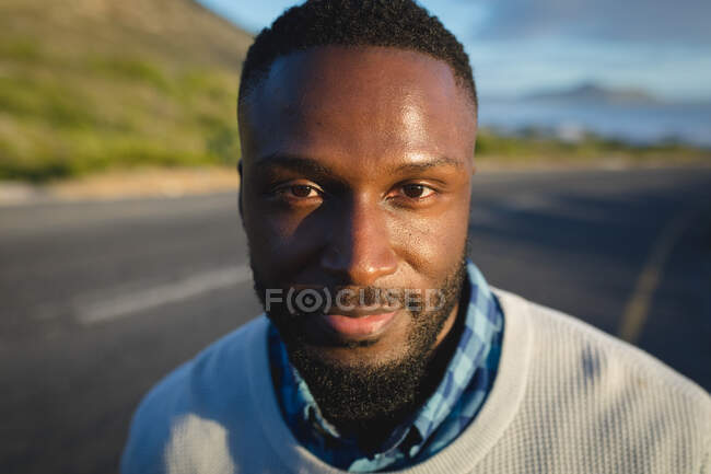 Porträt eines Afroamerikaners, der am Straßenrand steht, in die Kamera schaut und lächelt. Sommerreisen auf einer Landstraße an der Küste. — Stockfoto