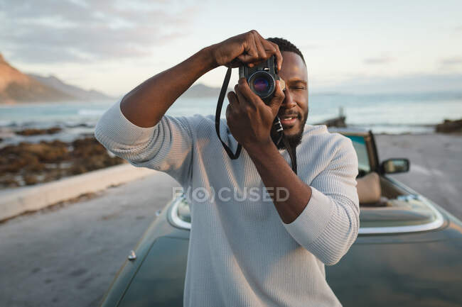 Homem afro-americano em pé junto ao carro conversível e tirando fotos com câmera. viagem de estrada de verão em uma estrada rural pela costa. — Fotografia de Stock